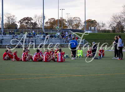 A group of young soccer players in red uniforms sit on a green field in the foreground, while spectators watch from bleachers in the background.