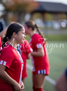 A young girl in a red soccer jersey stands on a green field in the foreground, with another player and blurred background elements behind her.