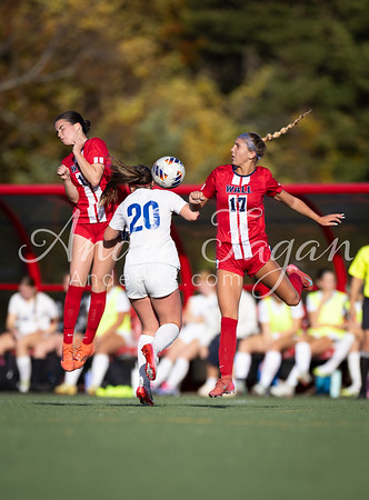 Three female soccer players in red and white uniforms jump to head a soccer ball on a green field, with spectators visible in the blurred background.