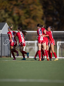 A group of female soccer players in red and white uniforms celebrate on a green field with a blurred background of trees and a building.