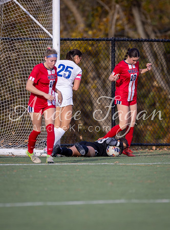 A soccer goalie lies on the ground with a ball at his feet while players in red and white uniforms stand nearby, with a fence and trees in the background.