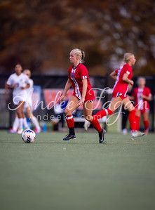 A female soccer player in a red uniform runs with a soccer ball on a green field, with other players and blurred trees in the background.