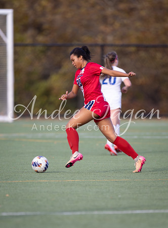 A female soccer player in a red uniform dribbles the ball on a green field with a blurred background of trees and a goalpost.