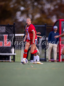 A female soccer player in a red and white uniform dribbles a soccer ball on a green field, with a blurred background of spectators and a fence.