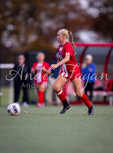 A female soccer player in a red and white uniform dribbles a ball on a green field, with blurred figures and a red bench in the background.