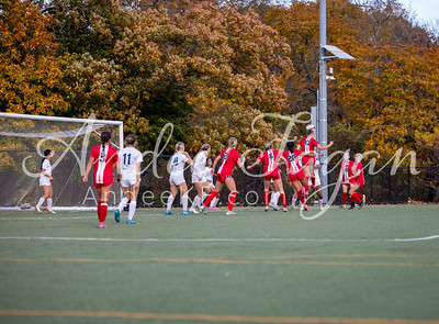 A soccer game is in progress on a green field with players in red and white uniforms, while a backdrop of autumn trees with colorful foliage frames the scene.