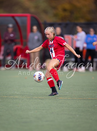 A young woman in a red soccer uniform kicks a ball on a green field, with blurred figures and a red bench in the background.