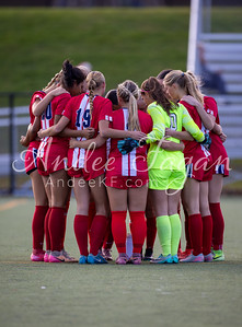 A group of female soccer players in red and white uniforms huddle together on a green field with a blurred background of bleachers and trees.