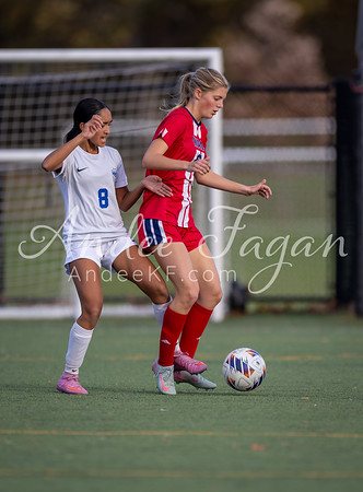 Two female soccer players in red and white uniforms battle for the ball on a green field with a blurred goal and fence in the background.