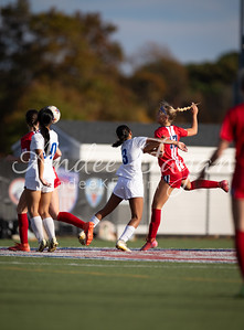 In the foreground, soccer players in red and white uniforms compete for the ball on a grassy field, while the background shows a blurred stadium with trees and a bright sky.