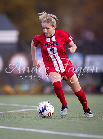 A female soccer player in a red and white uniform dribbles a ball on a green field with a blurred background of trees and buildings.