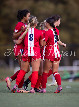 A group of young women in red soccer uniforms celebrate on a grassy field with trees in the blurred background.