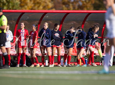 A group of young women in red and blue athletic wear stand on a green field in front of a red bench, with trees and a bright sky in the background.