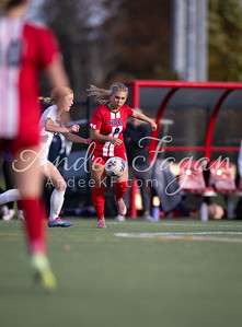A soccer player in a red uniform dribbles the ball on a green field with a blurred background of spectators and a red structure.