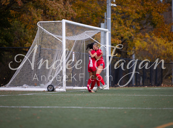 Two soccer players in red uniforms embrace in front of a goal, with autumn foliage visible in the background.