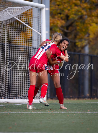 Two soccer players in red uniforms celebrate a goal in front of a net, with autumn foliage visible in the background.