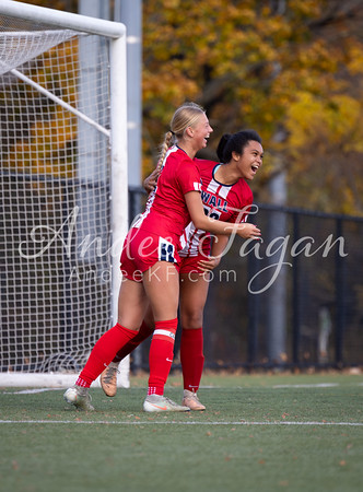 Two excited soccer players in red uniforms celebrate on a green field in front of a goal, with autumn trees in the background.