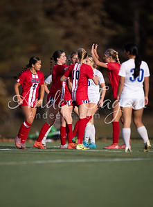 A group of young women in red and white soccer uniforms celebrate on a green field with trees in the blurred background.