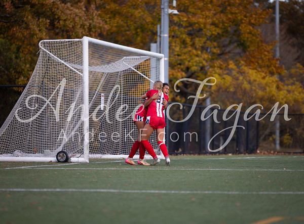 Two soccer players in red and white uniforms embrace near a goal on a field, with autumn trees in the blurred background.