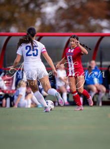 Two female soccer players in uniform are on a field with a blurred background of spectators and a red awning.