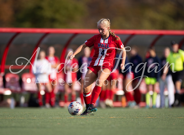 A female soccer player in a red uniform kicks a ball on a green field, with teammates and spectators blurred in the background.