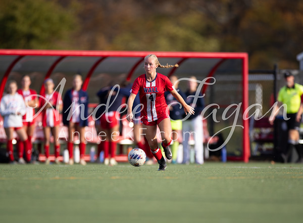 A young woman in a red soccer uniform dribbles a ball on a green field with a team on the sideline in the background.