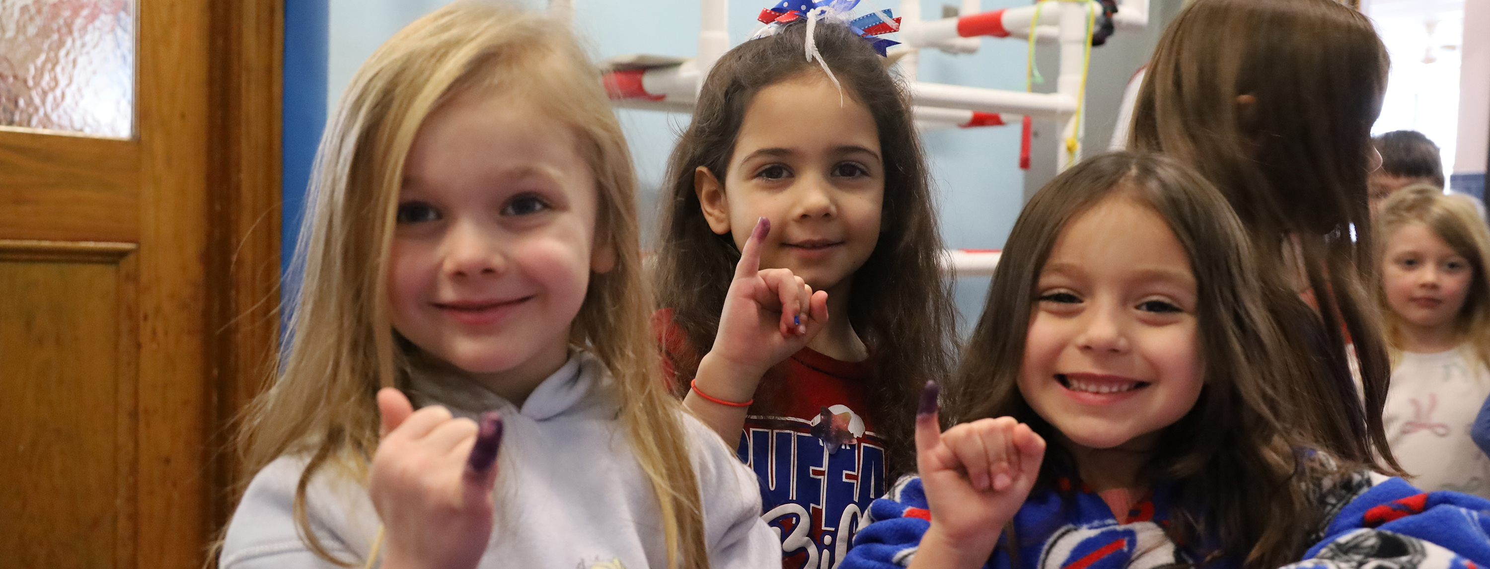 Three young girls with purple ink on their fingers smile at the camera in front of a classroom with colorful decorations.