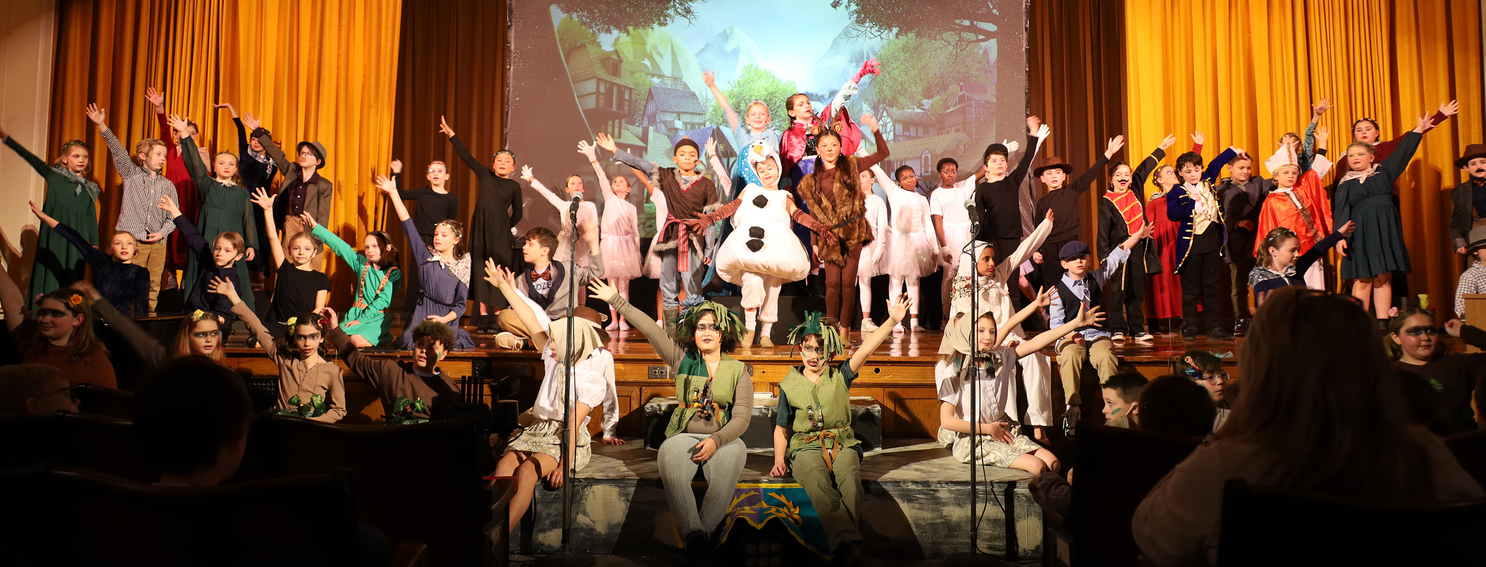 A group of children in costumes wave from a stage with a forest scene projected behind them, while an audience watches from the dark foreground.
