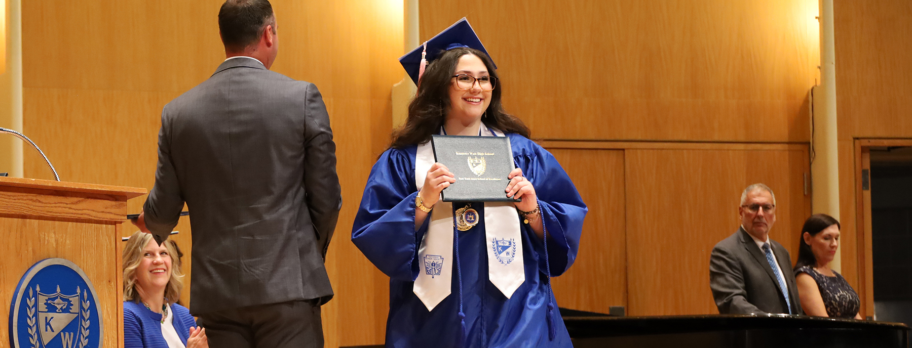 A graduate in a blue cap and gown holds her diploma in the foreground, while a man in a suit stands at a podium and other attendees watch in the background.