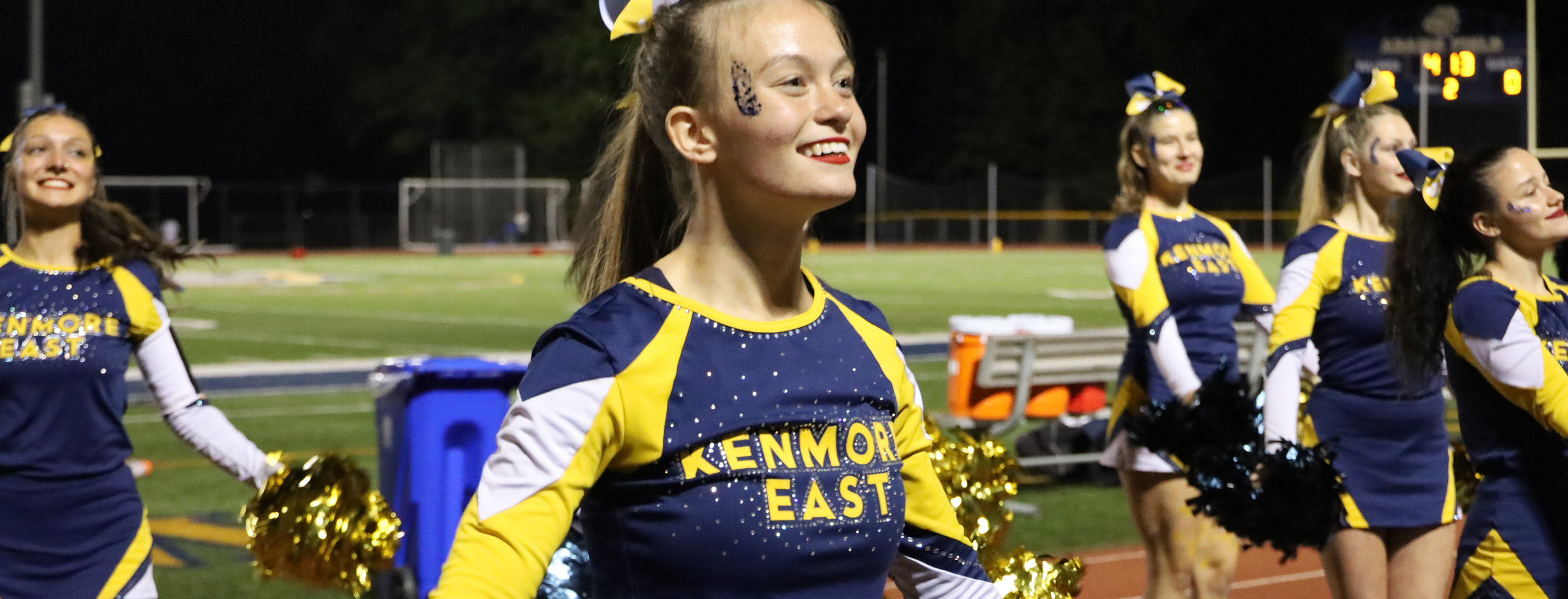 A group of smiling cheerleaders in navy and yellow uniforms with gold pom-poms stand on a track in front of a dimly lit football field.