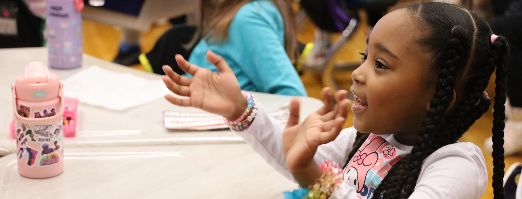 A young girl with braided hair and colorful bracelets on her wrist gestures with her hands while sitting at a table with a pink water bottle in the foreground and other children in the blurred background.