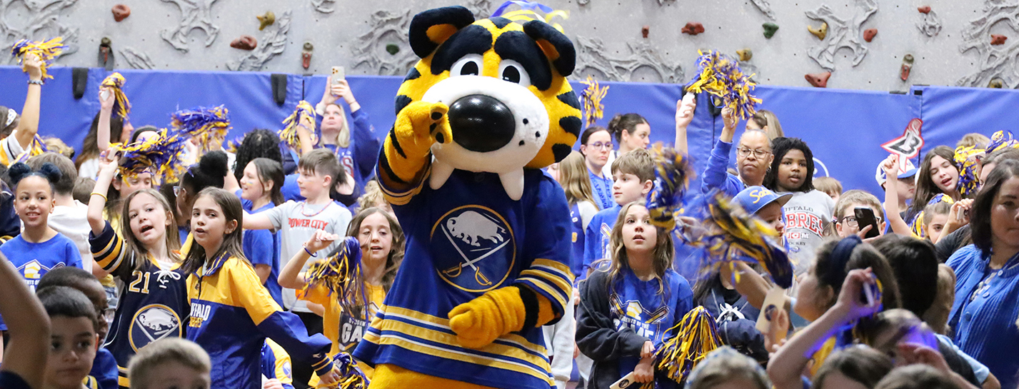 A mascot in a blue and gold jersey stands in front of a crowd of cheering children in a gymnasium with a climbing wall in the background.