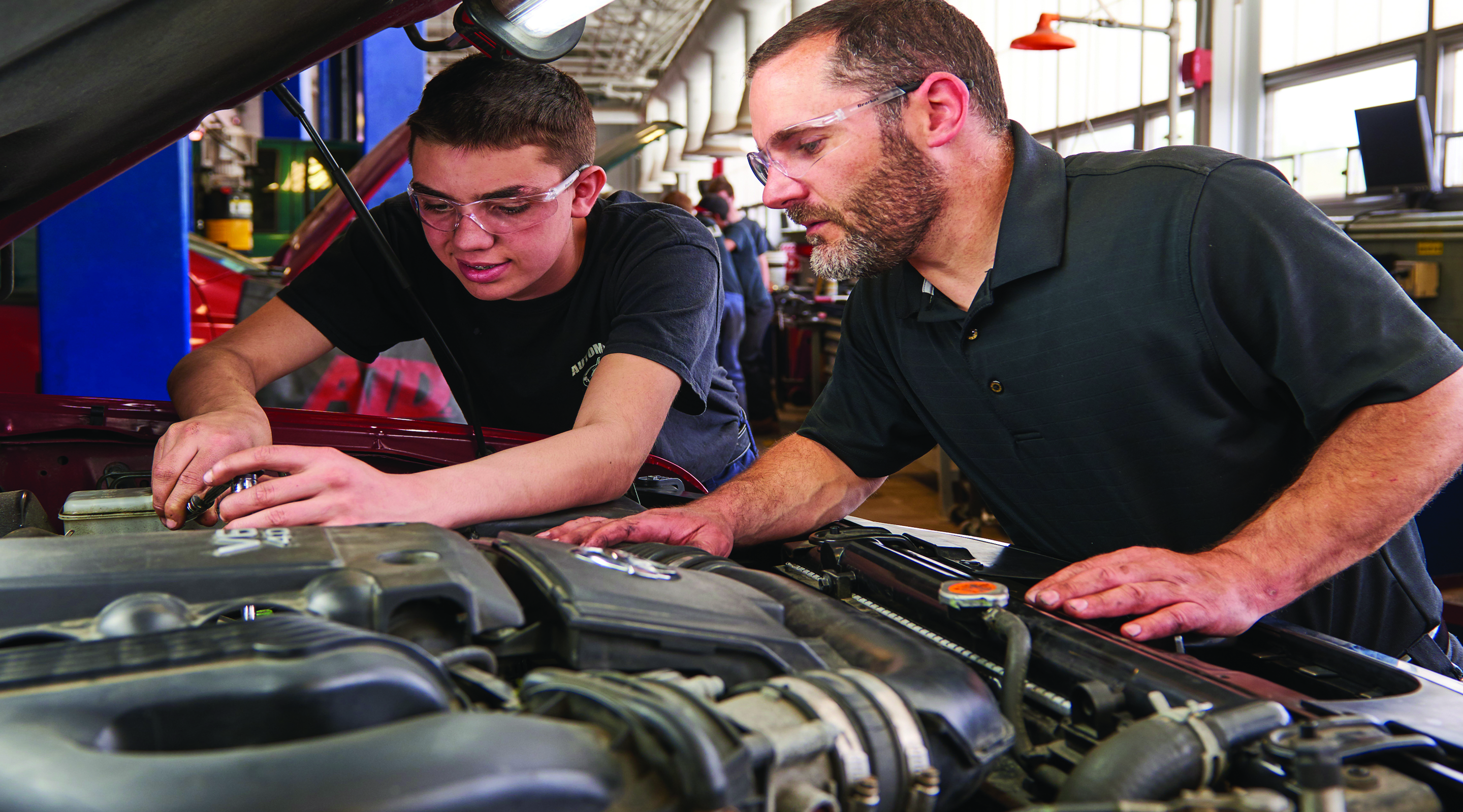 A male student and a male teacher work on a car engine in a brightly lit workshop.