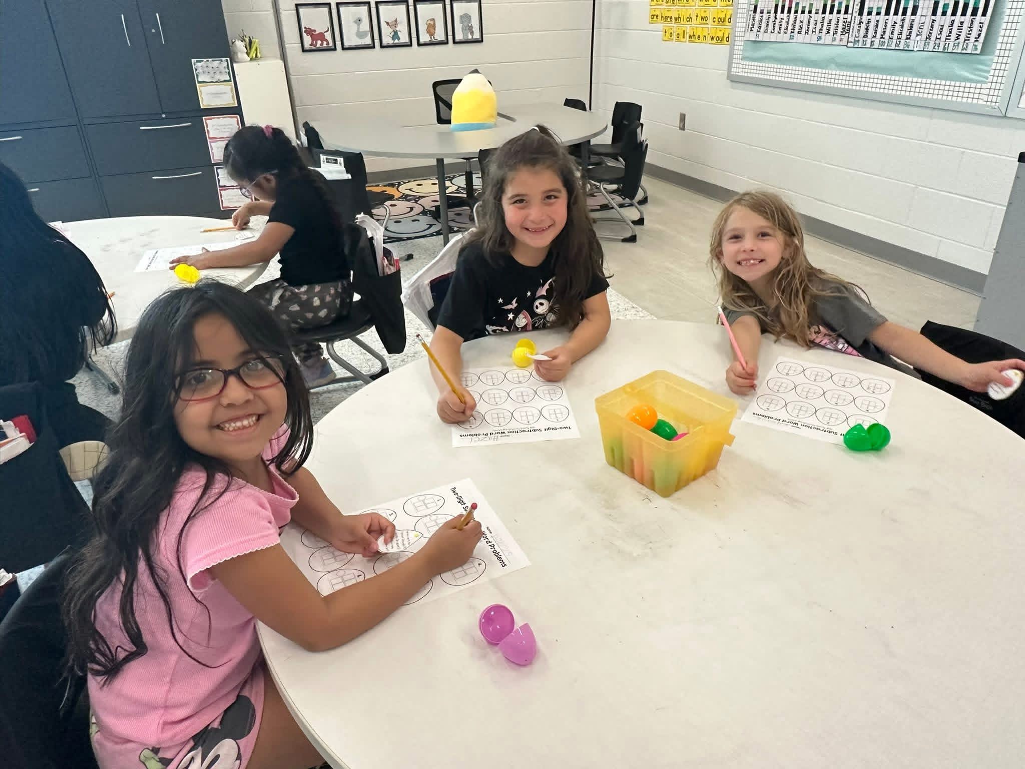 Three girls complete a plastic egg math activity at a classroom table.