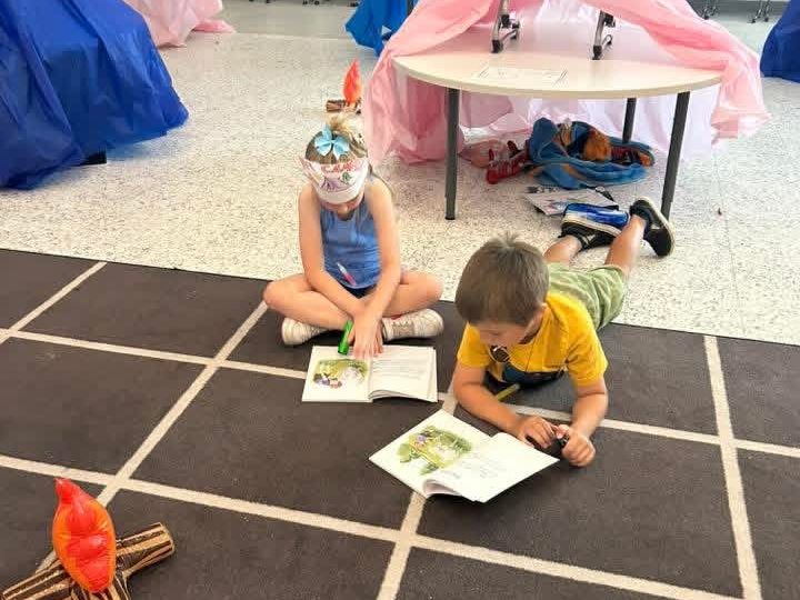 Two students read books on the floor near a pretend campfire and pretend tents.