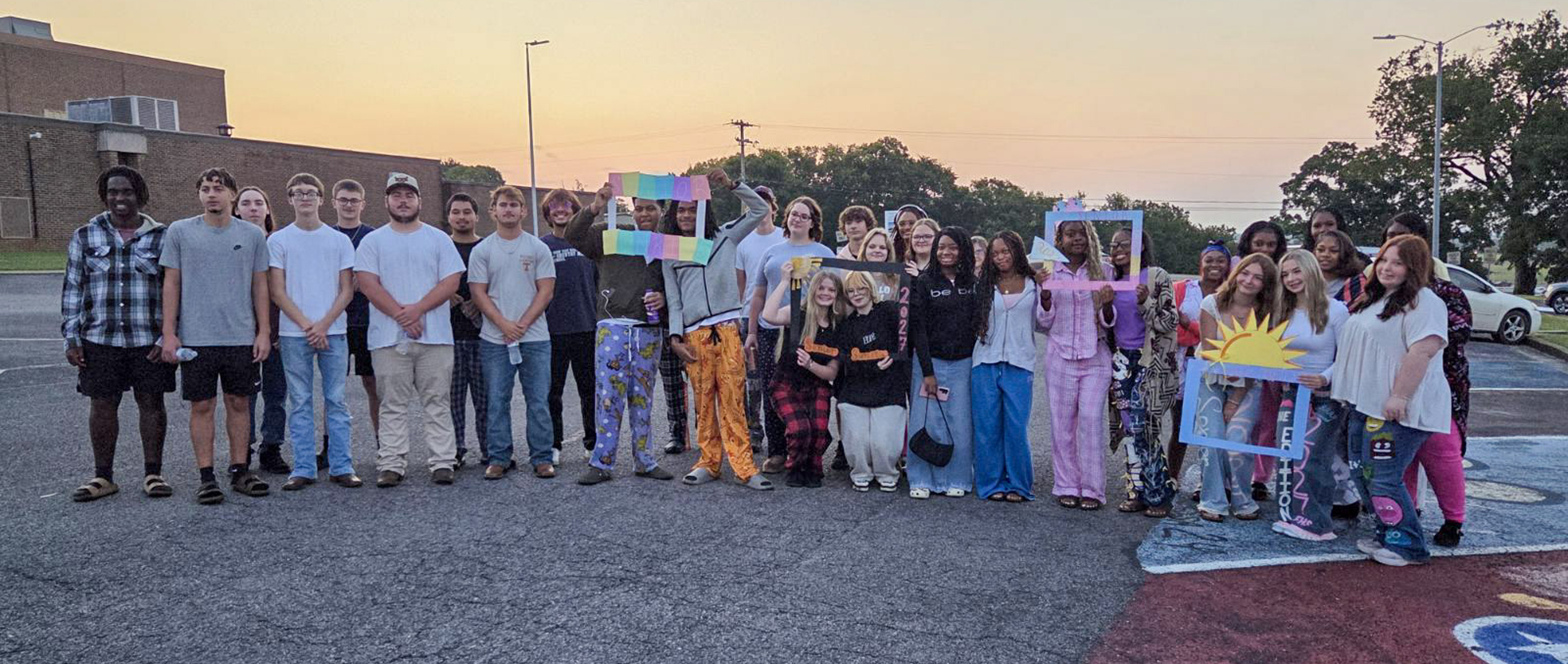 A group of young people in pajamas pose with decorative frames in front of a building at dusk.