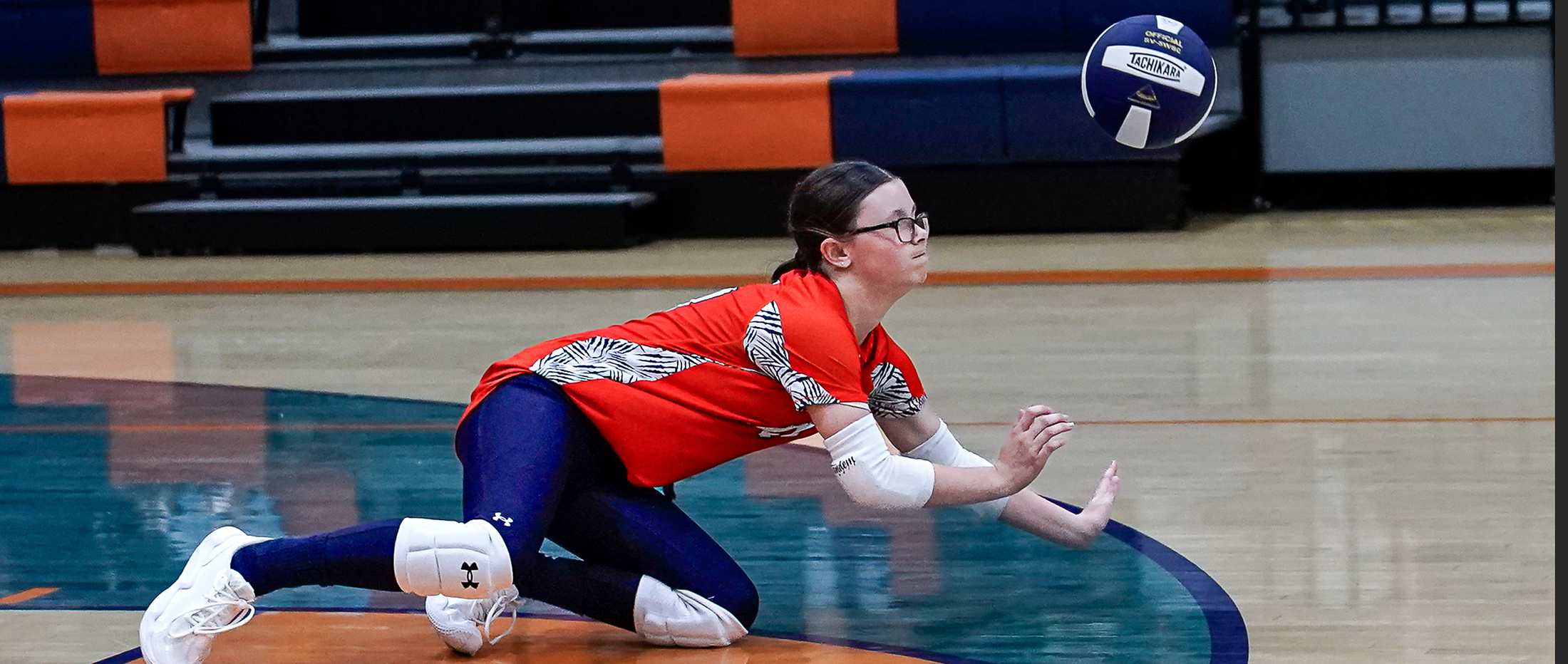 A volleyball player in an orange jersey dives to receive a ball on a wooden court, with bleachers in the background.