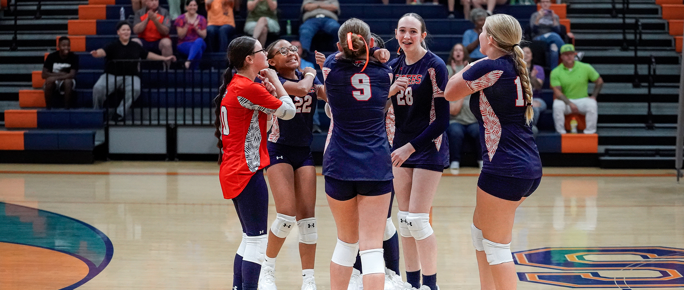 A group of young female volleyball players in uniform huddle together on a court with spectators in the bleachers behind them.