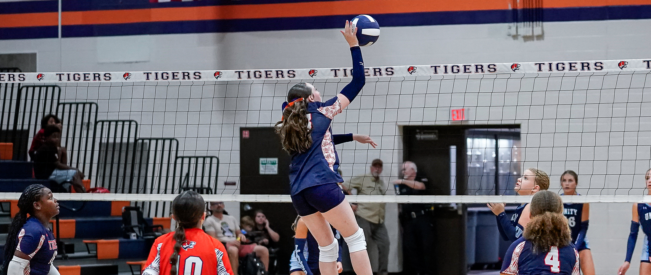 A volleyball player in the foreground jumps to hit the ball over the net, with spectators and a gymnasium in the background.