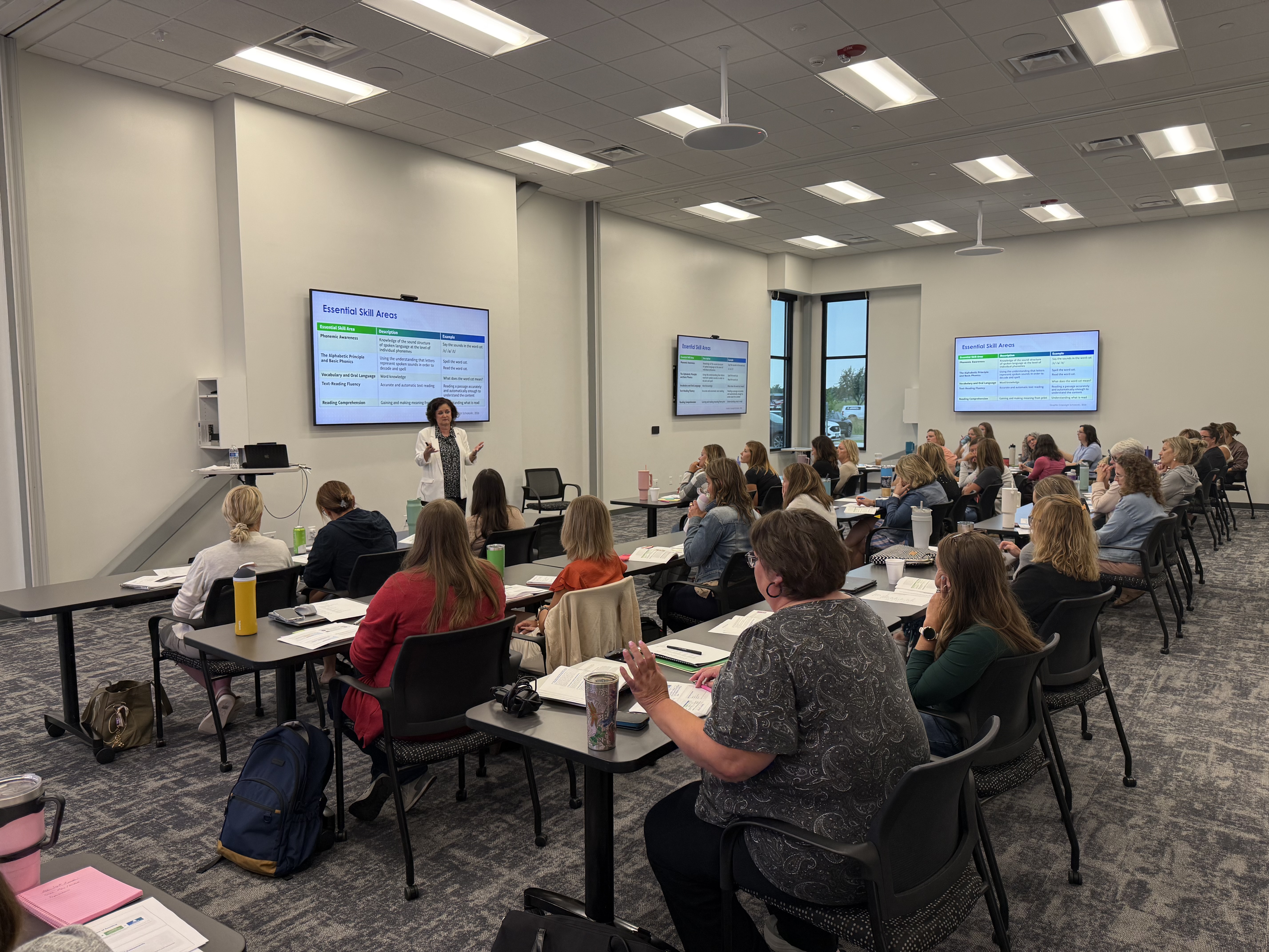 A presenter stands before a large screen in a classroom filled with seated attendees, with tables and chairs arranged in rows.