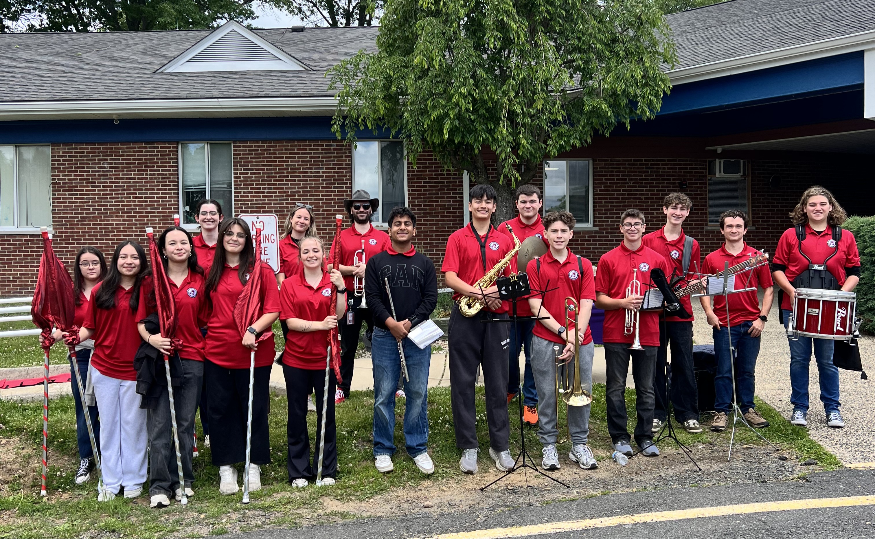 A group of young musicians in red shirts stand with their instruments and flags in front of a brick building.