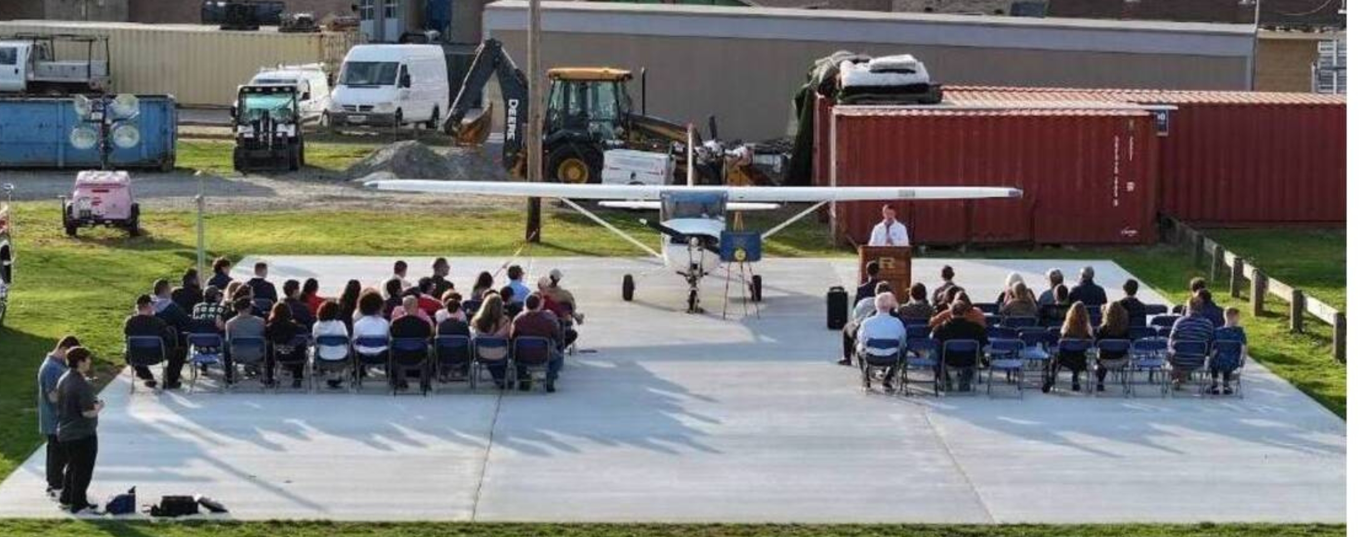 A small airplane sits on a tarmac in front of a seated audience of the Aviation Program with construction vehicles and shipping containers in the background.