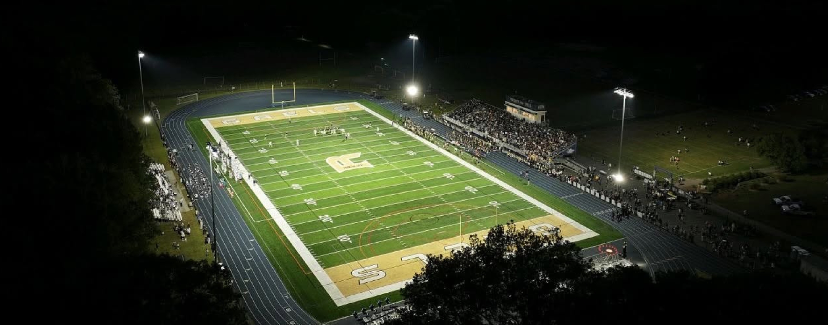 A football game is underway on a brightly lit field with spectators in the stands, set against a dark, nighttime sky.