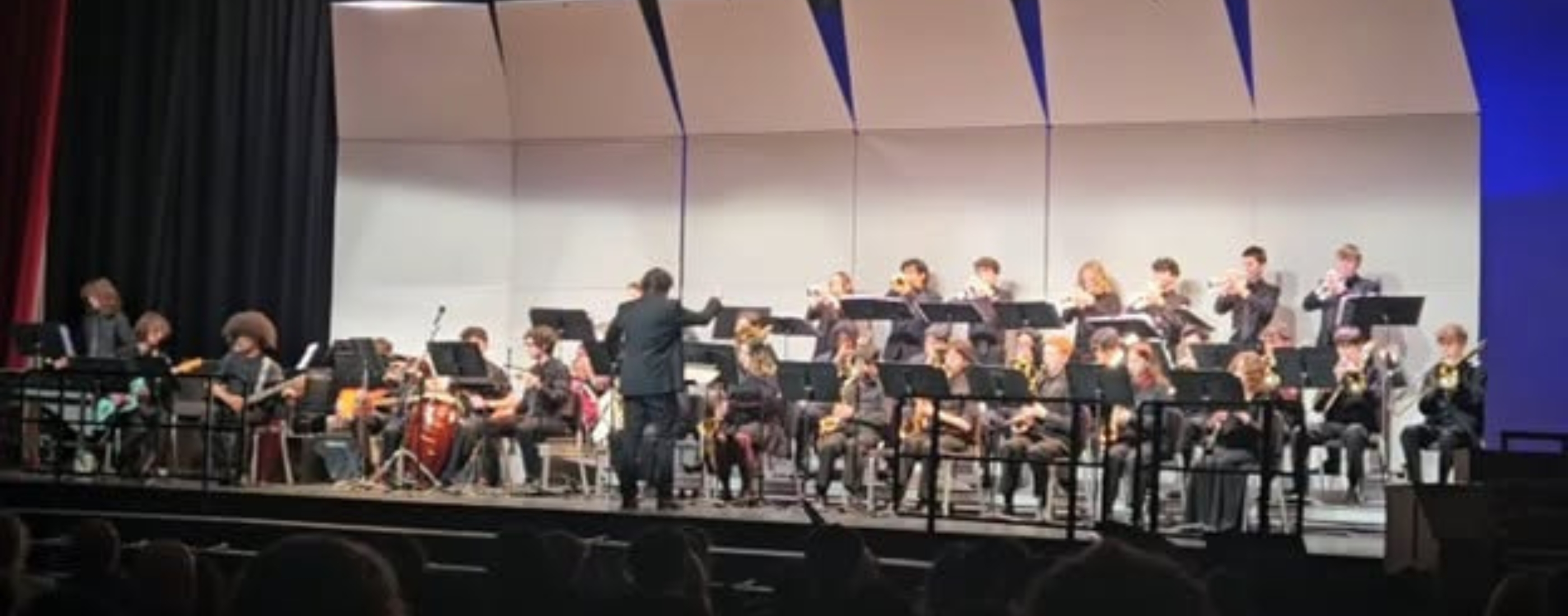 A conductor leads a band playing instruments on a stage with a patterned backdrop.