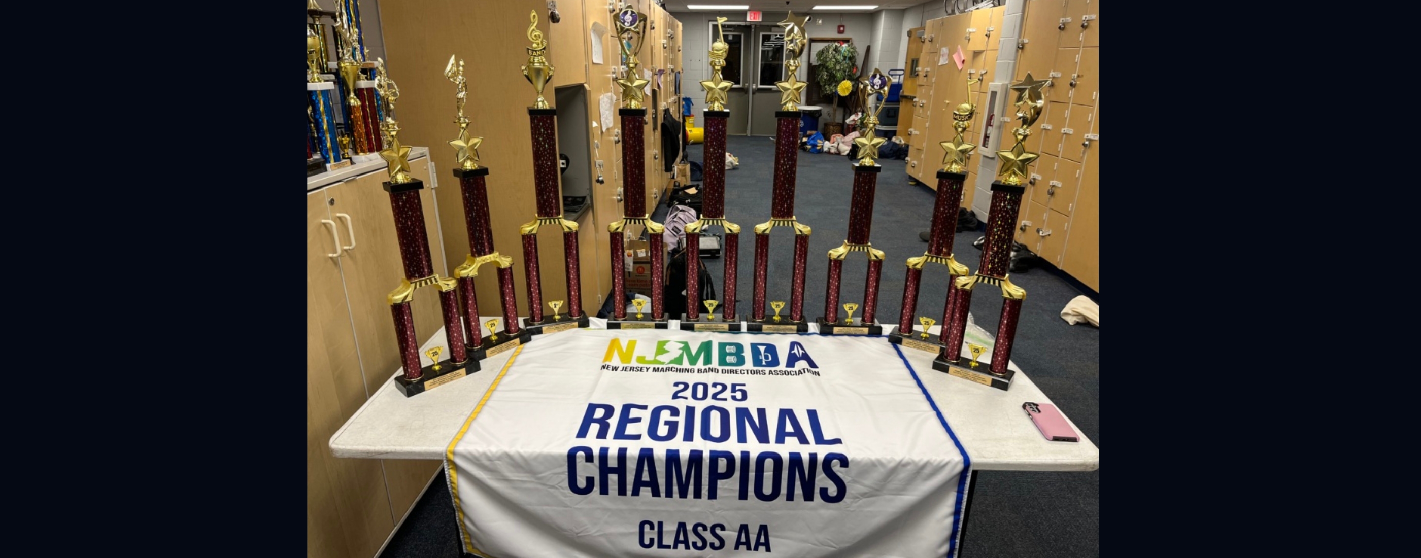 A collection of trophies sits on a table draped with a banner proclaiming "2025 Regional Champions Class AA" in front of a wall of lockers.