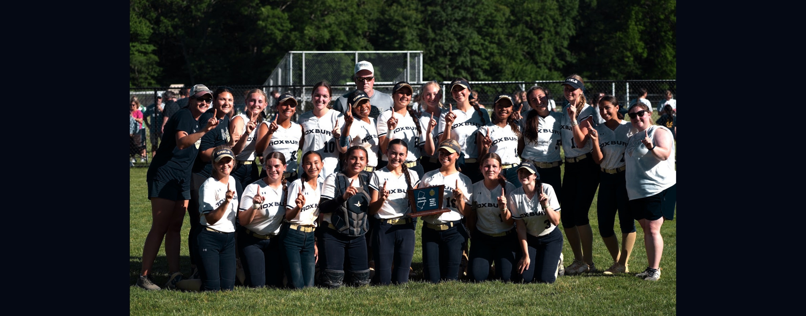 A group of young women in softball uniforms pose for a photo on a grassy field with trees and a fence in the background.