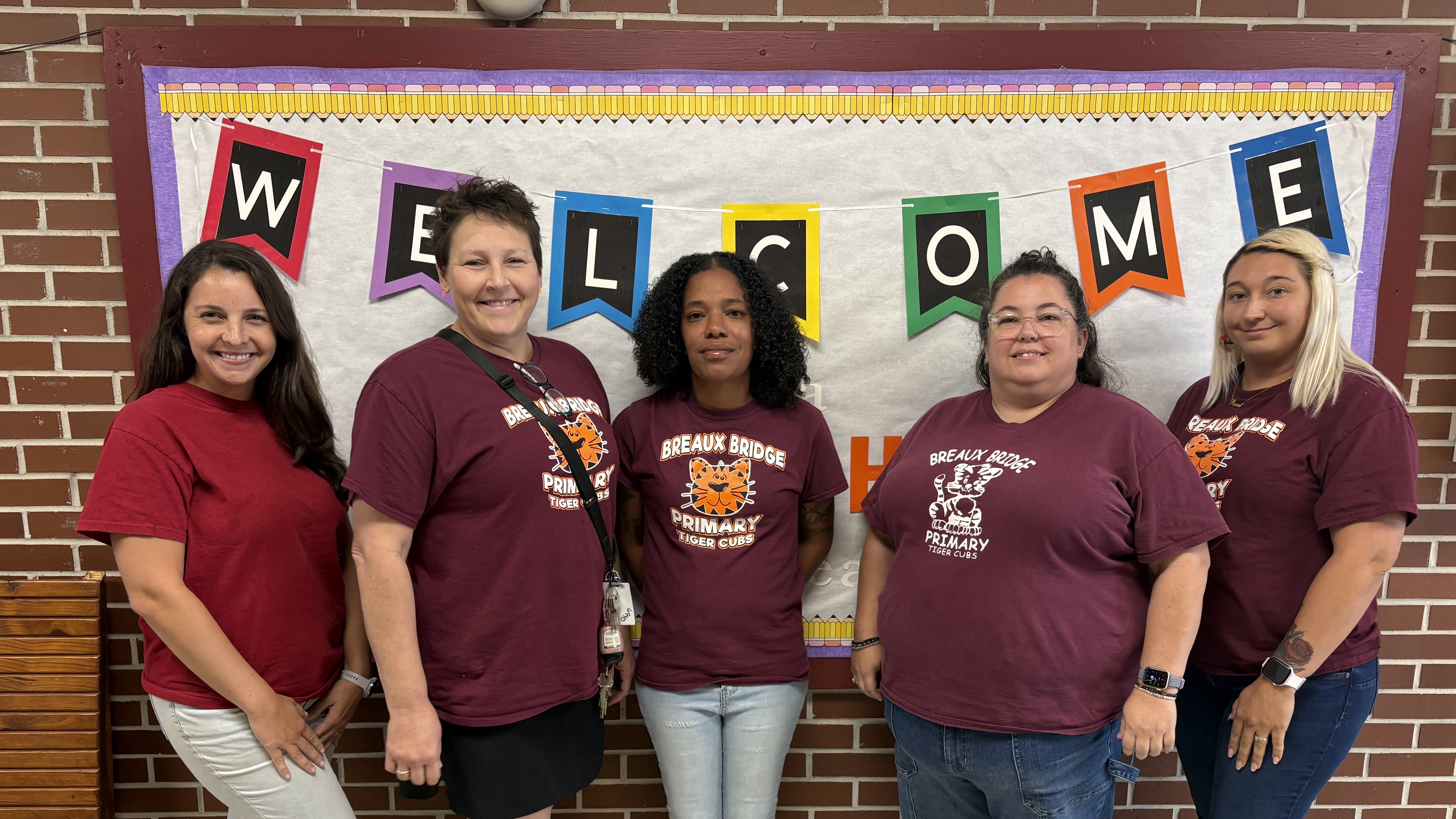 Five women wearing maroon t-shirts stand in the foreground in front of a "WELCOME" banner and a brick wall.