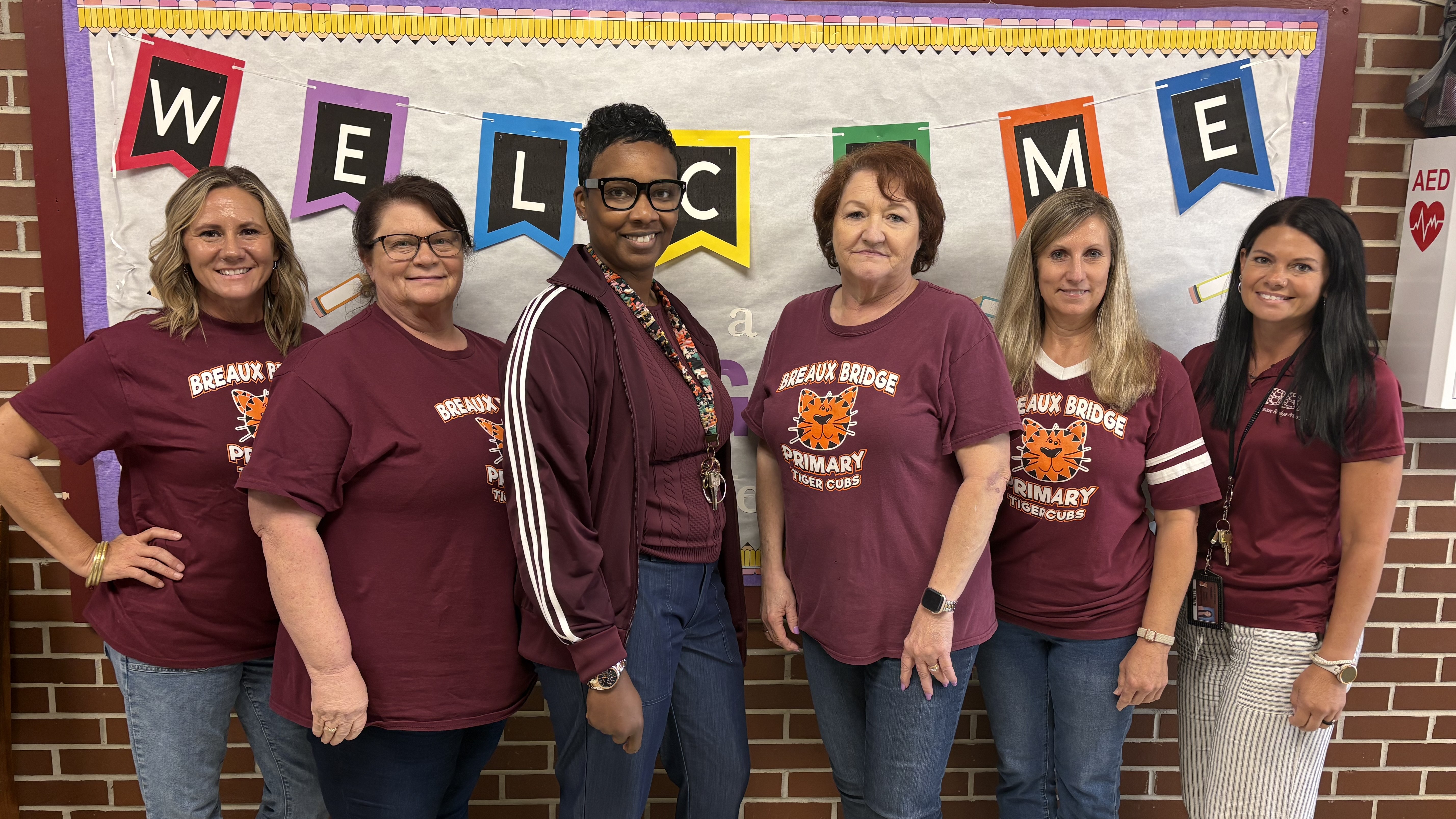 Six women wearing maroon shirts stand in the foreground in front of a "WELCOME" banner and a brick wall.