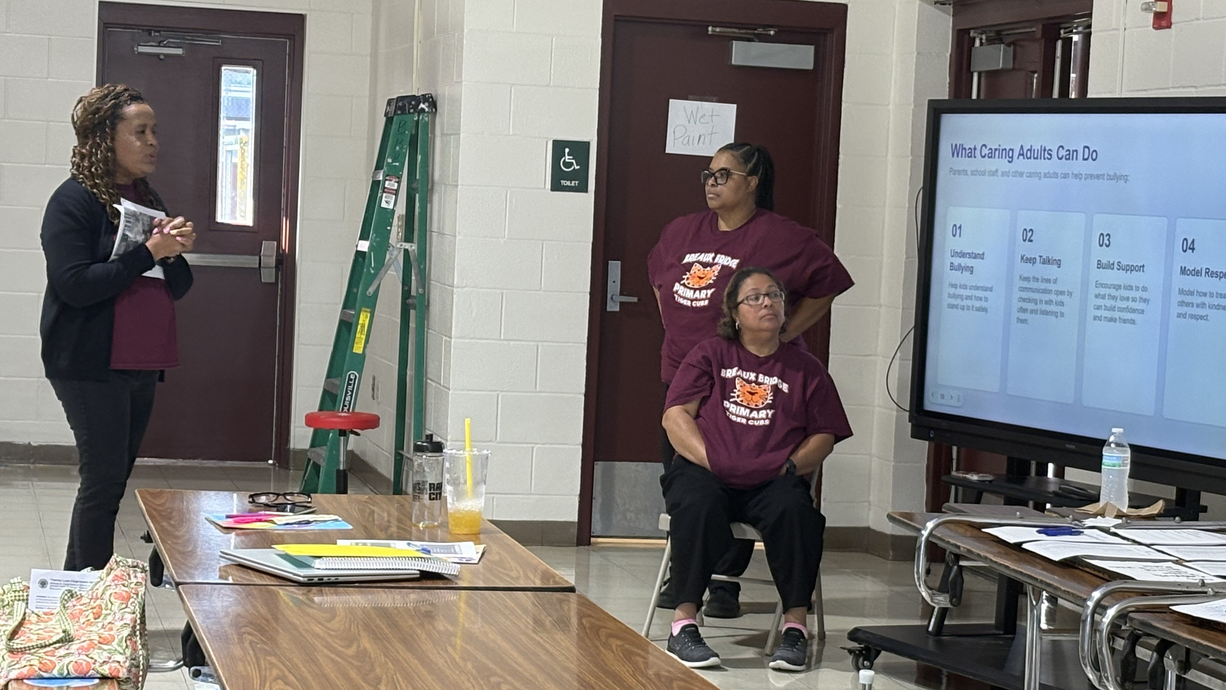 A woman stands speaking in the foreground next to a table, while two women sit near a presentation screen in a classroom background.
