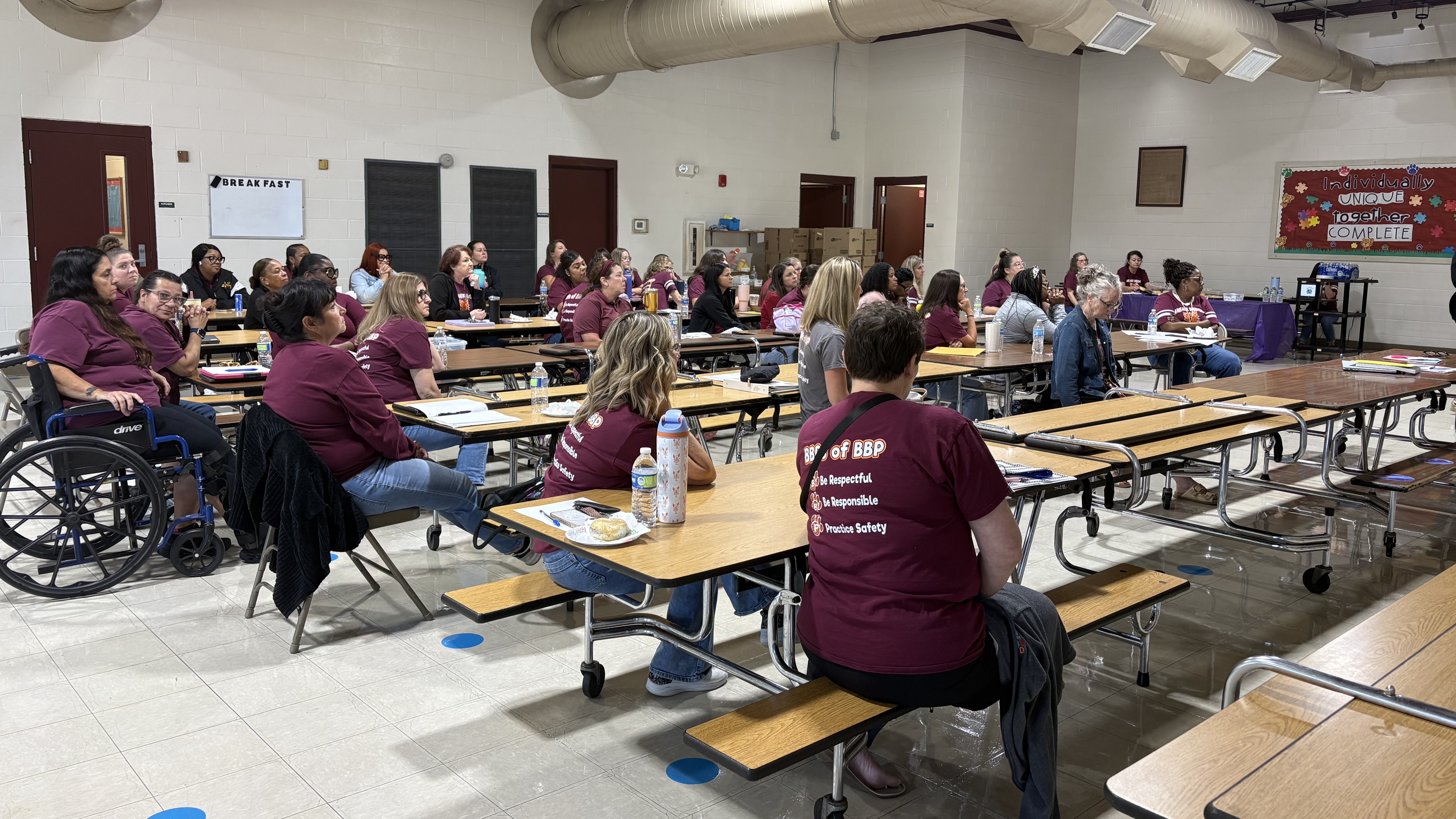 A group of people in maroon shirts sit at long tables in a large room with white walls and a colorful mural in the background.